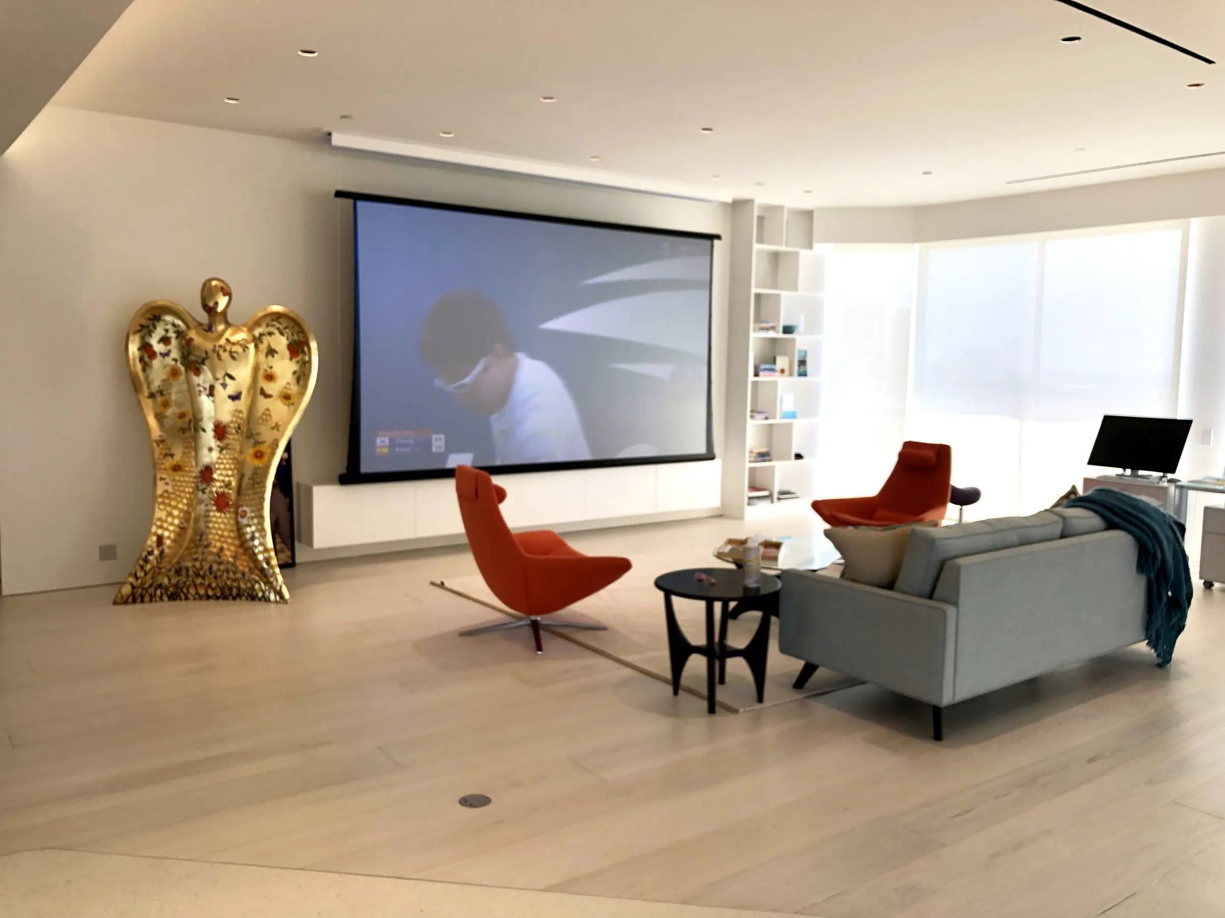 Modern living room with orange lounge chairs, a gold sculpture, and a projection screen showing a film