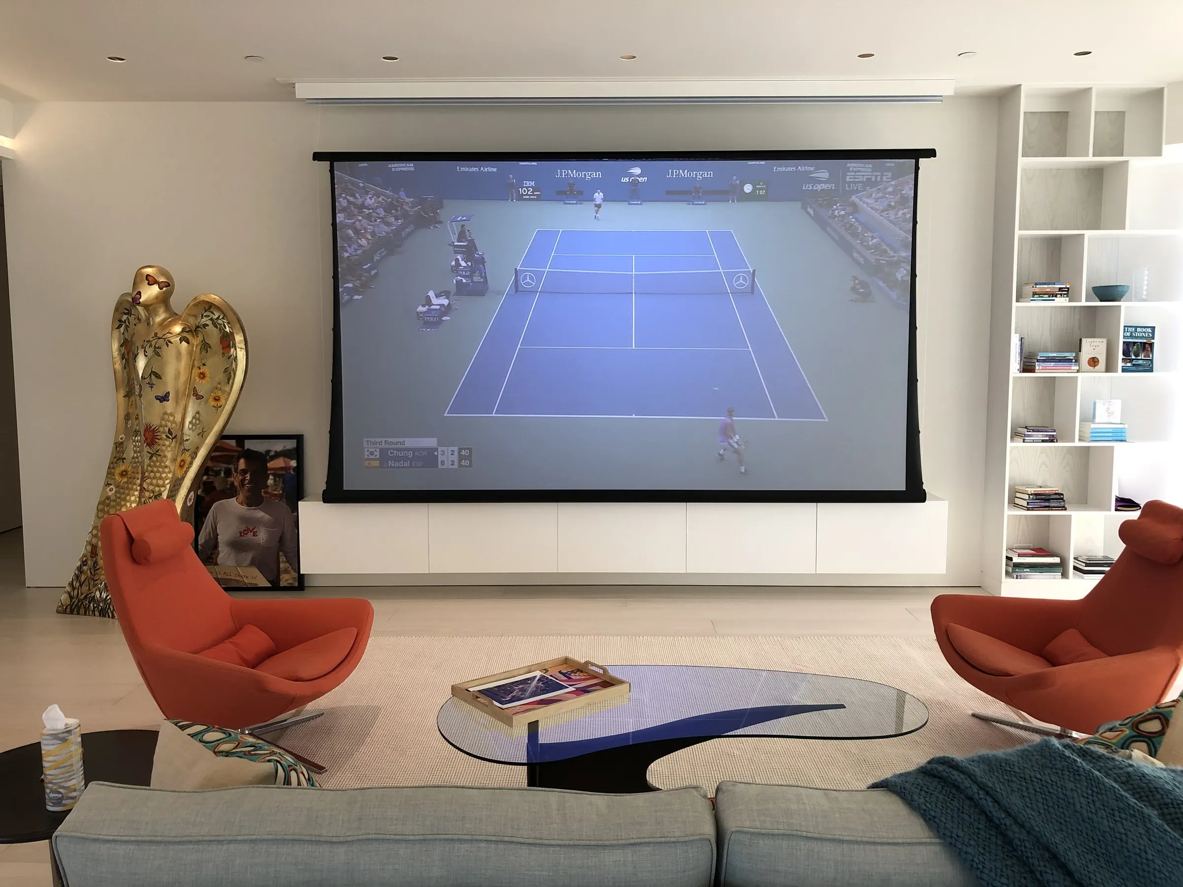 Modern living room with orange lounge chairs and a projection screen showing a tennis match