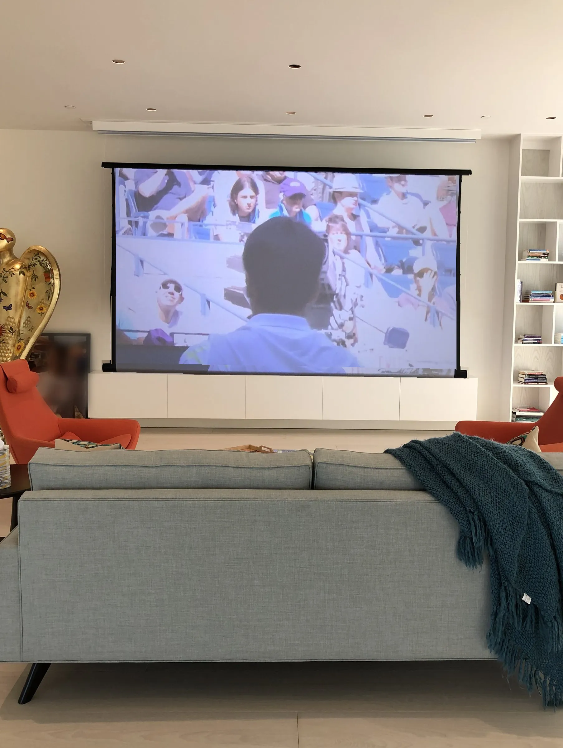 The same living room with a motorized projection screen lowered to the top of the cabinet, covering the art wall, a film playing