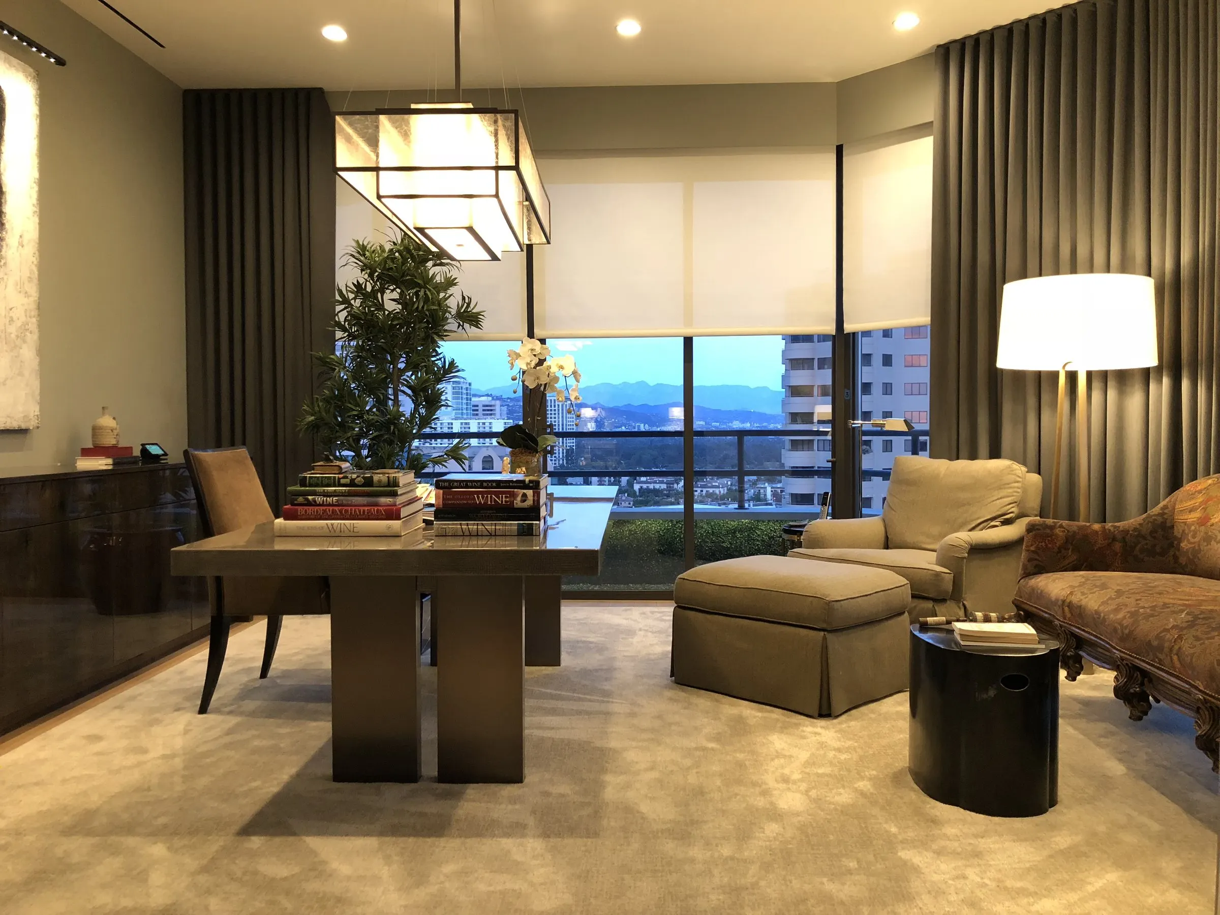 Home office with a roller shade lowered over city-view windows and a lamp-lit desk